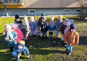 przedszkolaki stoją na łące pod blokiem wokół przebiśniegów