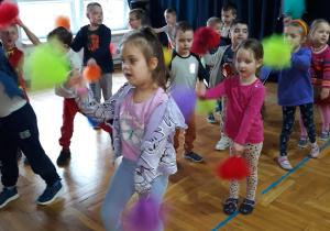 przedszkolaki tańczą ustawione w szeregach z kolorowymi pomponami, machając jednym z nich w górze