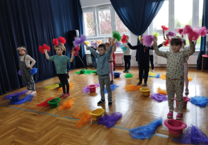 przedszkolaki stoją w szeregach, przy kolorowych kapeluszach i chustach, z uniesionymi rękami w górze z kolorowymi chustami zamocowanymi na gumkach