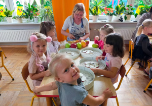 dzieci siedzą przy stoliku i układają jabłka na suszarkach, które kroi mama