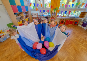 chłopiec w urodzinowej czapce siedzi wśród balonów na chuście, którą trzymają dziewczynki