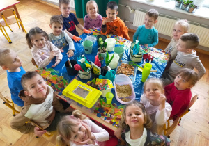 przedszkolaki siedzą przy stoliku, na kórym stoją sadzonki, pojemniki i narzędzia ogrodnicze nasiona,