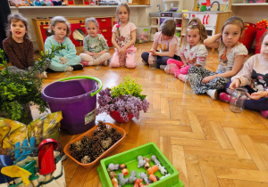 dzieci siedzą w półkolu ze słoikami wokół zgromadzonych gałązek, kwiatów i narzędzi potrzebnych do sadzenia