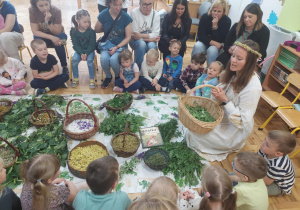 mamy z dziećmi siedzą wokół zgromadzonych ziół i prowadzącej ubranej w białą sukienkę i wianek
