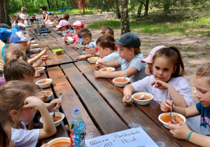 dzieci jedzą zupę przy drewnanych stołach rozstawionych na powietrzu