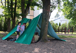dzieci pod namiotami wykonanymi z materiału zawieszonego na sznurku rozciągniętym pomiędzy drzewami