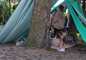 dzieci pod namiotami wykonanymi z materiału zawieszonego na sznurku rozciągniętym pomiędzy drzewami