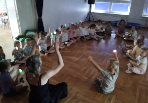 dzieci z nauczycielką siedzą w kole i bawią się światłem z latarek w zaciemnionej sali