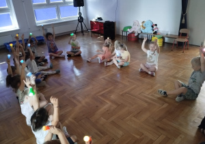 dzieci z nauczycielką siedzą w kole i bawią się światłem z latarek w zaciemnionej sali