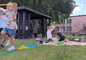 dzieci w ogrodzie bawią się rurkami antystresowymi siedząc na macie na trawie