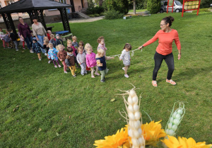 przedszkolaki idą w korowodzie za nauczycielką po ogrodzie przedszkolnym