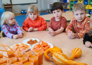 dzieci ubrane na pomrańczowo siedzą przy stoliku i próbują sok pomarańczowo-truskawkowy