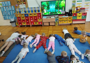 pani psycholog z dziećmi wykonuje ćwiczenie z jogi - leżenie na brzuchu z wyciągniętymi rękami na głową