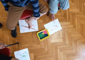 przedszkolaki próbują malować pędzelkiem trzymanym przez palce nogi