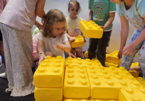 dzieci wznoszą budowlę z żółtych, plastikowych, dużych klocków