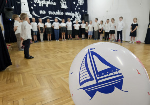 starszaki stoją w półkolu i śpiewają na tle balonu z niebieską łódką