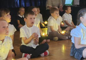 przedszkolaki śpiewają siedząc w ciemności i trzymając w rękach zapalone lampki