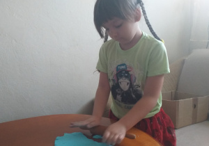 dziewczynka w warkoczach stoi przy stole i wałkuje wałkiem niebieskie ciasto