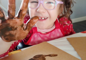 dziewczynka w okularach i kitkach pokazuje dłoń umalowaną na brązowo nad brązowym odciskiem dłoni na kartce