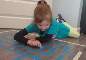 dziewczynka leży na podłodze ze zgiętymi nogami i przesuwa niebieską nakrętkę po naklejonym na podłogę labiryncie