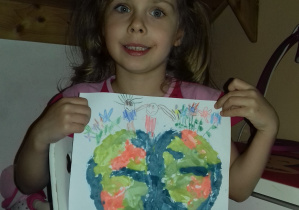 dziewczynka w różowej bluzce pokazuje pracę wykonaną farbami rosnącymi: planeta ziemia a na niej narysowana para wśród kwiatów