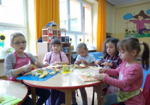 dziewczynki przy stoliku siedzą w fartuszkach i kroją owoce na deskach na sałatkę owocową