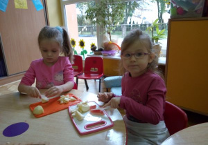 dwie dziewczynki siedzą przy stoliku i kroją na desce owoce