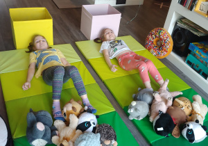 siostry leżą na zielonych materacach i nogami przenoszą zabawki do pudła ustawionego za głową