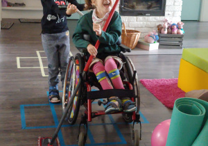 dziewczynka prowadzi wózek, na którym siedzi jej siostra i trzyma mopa do podłogi