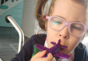 dziewczynka w okularach dmucha w gwizdek w kształcie fioletowego samolotu ze smigłem