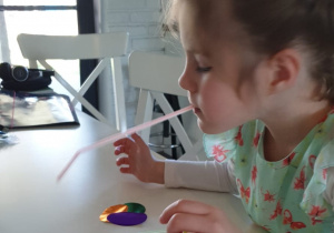 dziewczynka słomką umieszczoną w ustach próbuje podnosić kolorowe, papierowe kółka