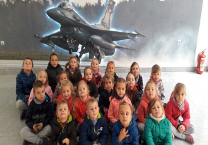 dzieci z gr VI siedzą w grupie przed ścianą z namalowanym samolotem wojskowym