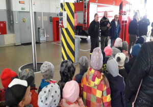 dzieci stoją w hangarze przed wozem strażackim i strażakami