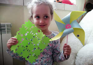 dziewczynka w związanych lokach pokazuje zieloną serwetkę wyciętą z papieru oraz niebiesko - żółty wiatrak zamocowany na słomce