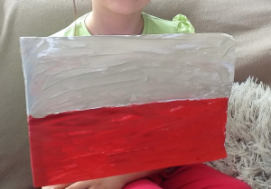 dziewczynka siedzi na łóżku i pokazuje dużą flagę Polski pomalowaną farbami plakatowymi zamocowaną na patyku