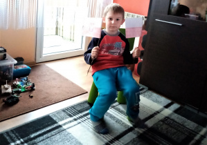 w domu chłopiec siedzi na środku pokoju, na krześle a w rękach trzyma dwie flagi Polski