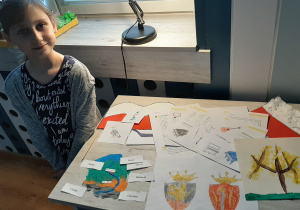 dziewczynka siedzi przy stole i pokazuje swoje prace plastyczne wykonywane podczas nauki zdalnej