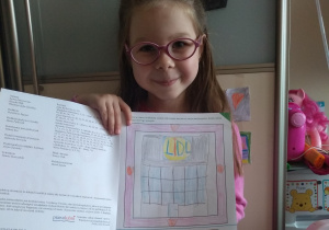 w domu, dziewczynka w rózowych okularach pokazuje książkę z wykonanym zadaniem