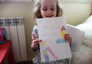 w domu, dziewczynka z lokami na głowie pokazuje kartkę z napisem "moja Zduńska Wola" i narysowanym słońcem, domem, huśtawką i zjeżdżalnią