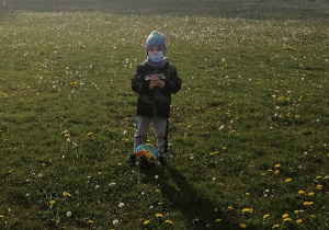 chłopiec stoi na łące w masce, z zabawką autobusem i piłką między nogami