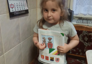 w domu stoi dziewczynka i trzyma książkę z wykonanym zadaniem: wydrukowany i wklejony herb Zduńskiej Woli i flagi miasta