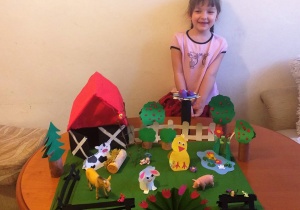 uśmiechnięta dziewczynka w różowej bluzce z kotami siedzi za stołem, na którym stoi makieta wiejskiej zagrody: budynki, płoty, drabina, drzewa wraz ze zwierzętami