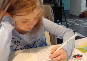dziewczynka siedzi w domu przy stoliku, podpiera głowę jedna ręką a drugą pisze ołówkiem w książce