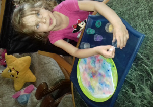 dziewczynka siedzi przy niskim stoliku i pipetą i farbą robi plamy na soli rozsypanej na zielonym talerzyku umieszczonym na dużej, granatowej tacy
