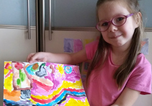 dziewczynka w okularach pokazuje swoją pracę plastyczną: namalowane morze kolorowymi falami i żółtym słońcem