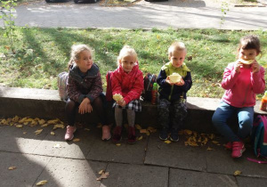 dzieci siedzą na dworze, na murku i jedzą bułki z serem