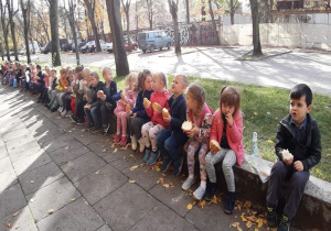 dzieci siedzą na dworze, na murku i jedzą bułki z serem
