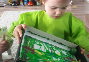 w domu, dziewczynka w zielonym fartuszku robi prace plastyczną: trzyma w rękach i porusza pudełkiem z naklejoną kartką w kolorowe plamy z farby i umieszczoną na niej kulką