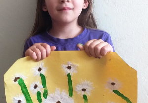 dziewczynka w fioletowej bluzce pokazuje wykonaną pracę plastyczną: na żółtej kartce dmuchawce z odciśniętej i pociętej rolki papierowej, brązowymi srodkami i namalowanymi zieloną farbą trawą i łodygami