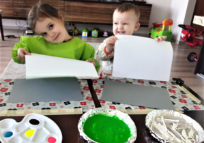 dzieci w domu, siedzą przy stole szykują się do pracy a przed nimi stoją talerze z farbami: zielona i białą